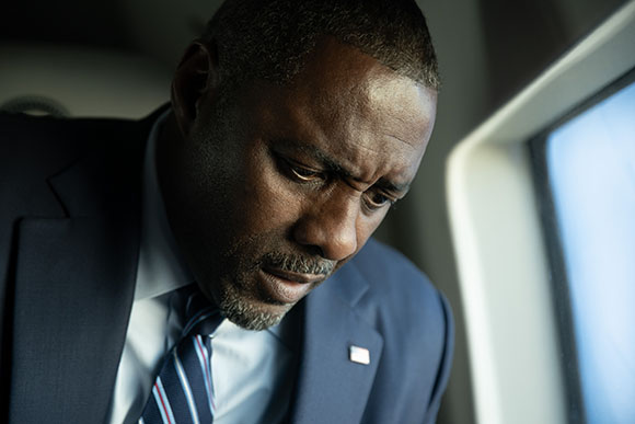 A man in a dark suit and striped tie looking downward with a serious, contemplative expression.