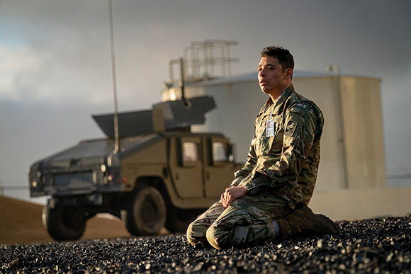 A soldier in camouflage fatigues sits on a gravel surface with a military vehicle in the background.
