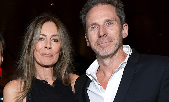 A man in a dark suit and white shirt stands next to a woman with long brown hair in a black top.