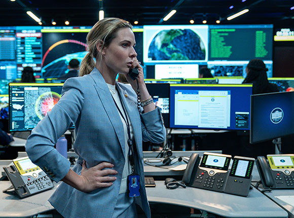 A woman in a light blue blazer speaks into a handset in a high-tech control room filled with monitors.