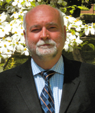 A bald man with a gray beard wearing a black suit, blue patterned shirt, and striped tie.