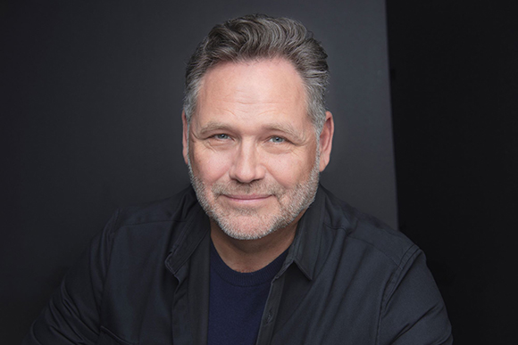 A middle-aged man with salt-and-pepper hair and a beard smiling against a dark background.