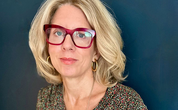 A blonde woman wearing red-rimmed glasses and gold hoop earrings against a dark blue background.