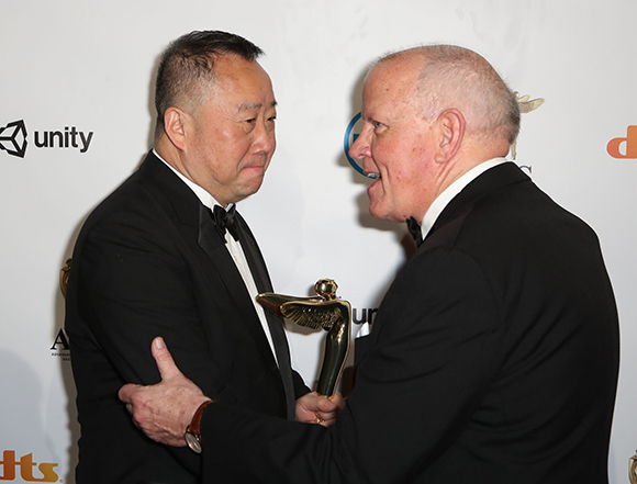 Two men in black tuxedos face each other while holding a gold trophy against a white backdrop.