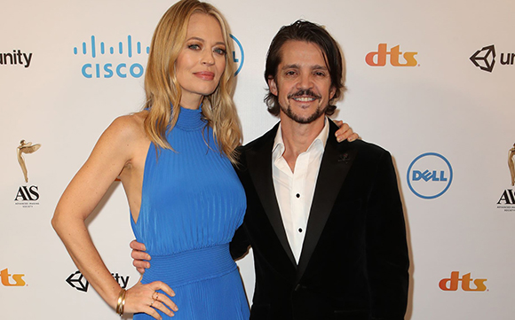 A blonde woman in a blue dress and a man in a black velvet blazer smiling at a red carpet event.