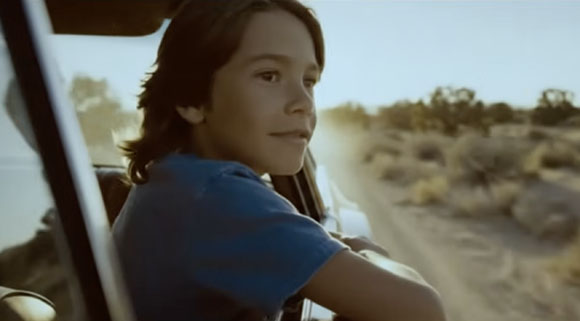 A young boy with brown hair looks out of a car window while traveling through a desert landscape.