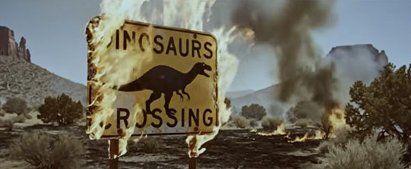 A yellow rectangular sign with a dinosaur silhouette is engulfed in flames in a desert landscape.