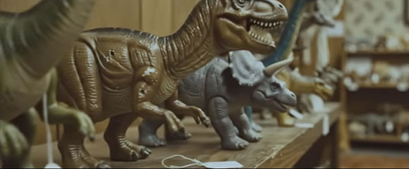 Several plastic dinosaur figurines, including a large brown T-Rex and a grey Triceratops, on a shelf.