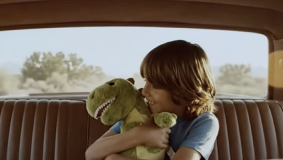 A young child with brown hair smiles while hugging a green plush dinosaur in a car seat.