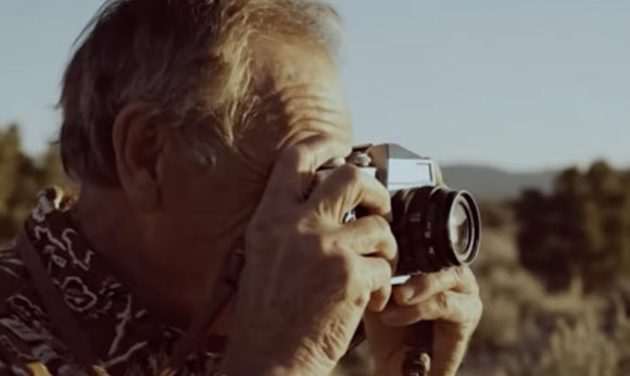 A man holds a vintage camera up to his eye to take a photo outdoors.