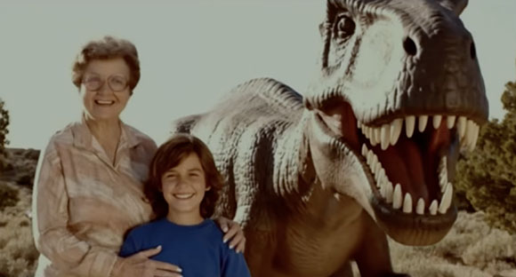 An elderly woman and a young boy smile while standing next to a large Tyrannosaurus Rex model.