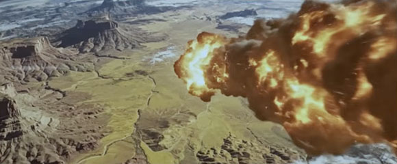 A large, bright orange and yellow fireball erupting over a vast, rocky desert canyon landscape.