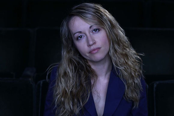 A woman with long, wavy blonde hair wearing a dark blue blazer looks toward the camera.