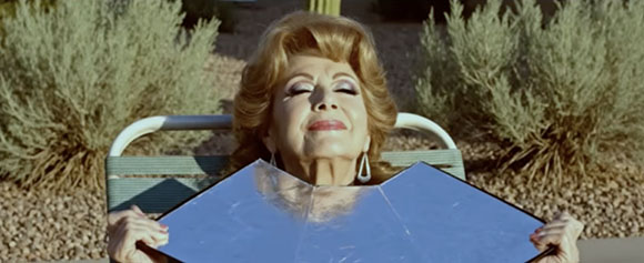 A woman with blonde hair reclines in a lounge chair, holding a large mirror in front of her chest.