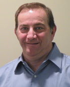 A smiling man with short brown hair wearing a light blue button-down shirt against a plain background.