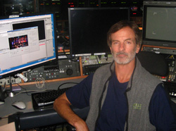 A man with graying hair and a beard sits in front of several computer monitors in a control room.