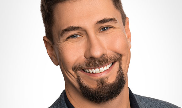 A close-up portrait of a smiling man with short brown hair and a dark goatee against a white background.