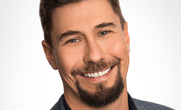 A close-up portrait of a smiling man with short brown hair and a goatee against a white background.