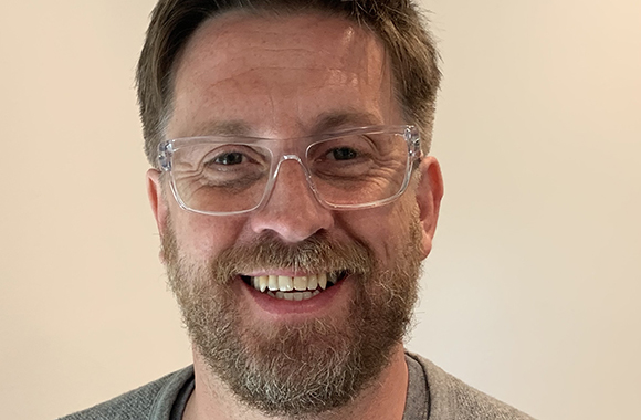A smiling man with brown hair, a beard, and clear-rimmed glasses looking at the camera.