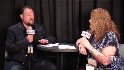 A man in a dark blazer speaks into a microphone while a woman with curly hair listens during an interview.