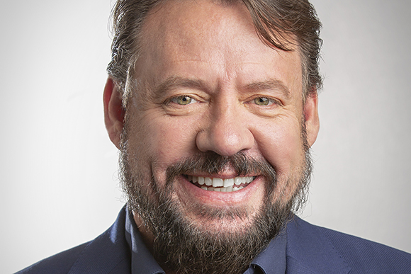 A smiling man with brown hair, green eyes, and a dark beard wearing a blue blazer.