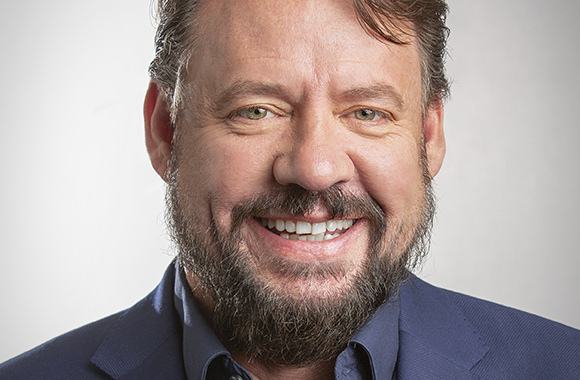 A smiling man with brown hair, green eyes, and a salt-and-pepper beard wearing a blue shirt and blazer.