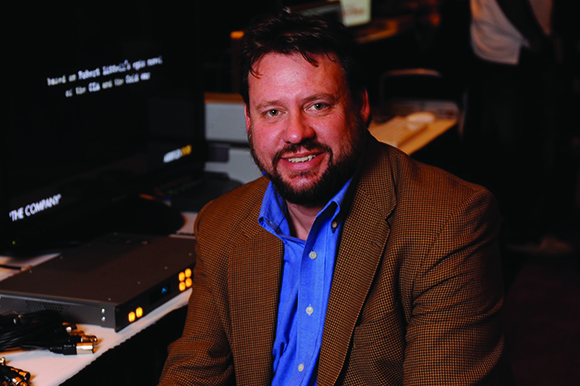 A man with a beard wearing a brown blazer and blue button-down shirt smiles toward the camera.
