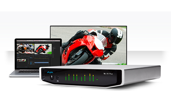 A silver AJA 4K Plus device in the foreground with a laptop and monitor showing motorcycle racing imagery.
