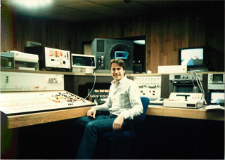 A man in a light shirt sits in a room surrounded by various monitors, control panels, and electronic devices.