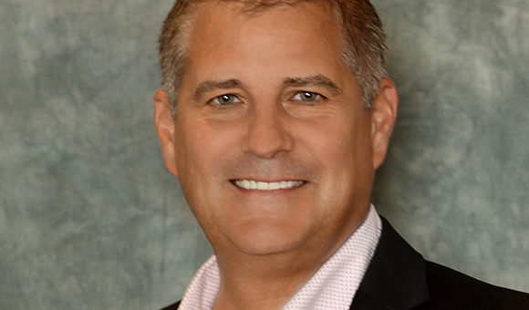 A smiling man with short graying hair wearing a light pink shirt and black jacket.