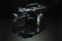 A professional black video camera with a large lens and a small remote control on a reflective black surface.