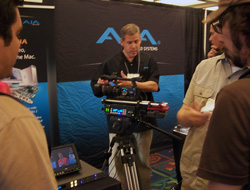 Man demonstrates a professional video camera on a tripod at a trade show booth with company branding in the background.