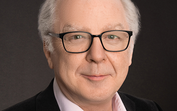 A man with white hair and black-rimmed glasses smiling slightly against a dark background.