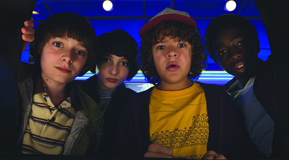 Four young boys looking forward with wide eyes and surprised expressions from inside a dark space.