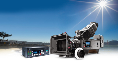 A professional cinema camera and a digital control unit set against a bright, sunny beach landscape.