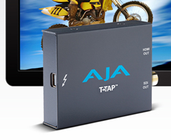 A dark gray AJA T-TAP device positioned in front of a monitor showing a yellow motocross motorcycle.