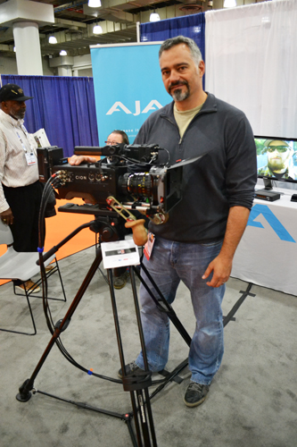 A man in a grey pullover and jeans stands beside a large professional cinema camera mounted on a tripod.