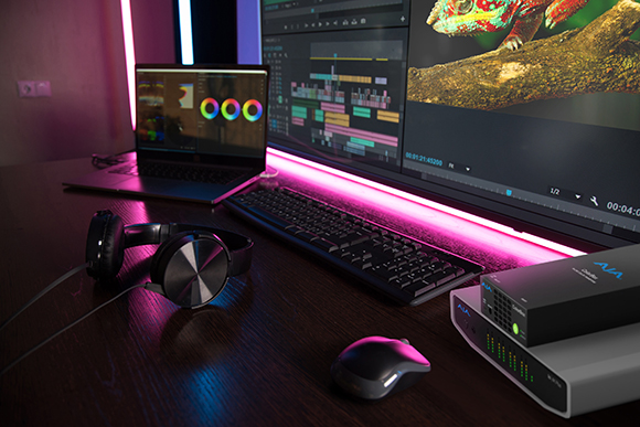 Video editing setup with dual monitors, laptop, keyboard, mouse, and headphones under pink neon lighting.