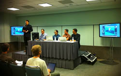 A panel of five men sits at a long table in a conference room with two large monitors displaying graphics.
