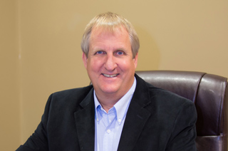 A smiling man with blond hair wearing a dark blazer and light blue button-down shirt.