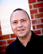 A man with short hair and a black shirt smiling in front of a red brick wall.