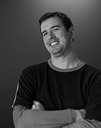 Black and white portrait of a smiling man with short hair, wearing a dark t-shirt and crossing his arms.