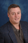A man with short brown hair wearing a dark blazer over a grey polo shirt against a blue background.