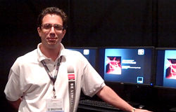 A man in a white polo shirt standing next to three computer monitors in a dark room.