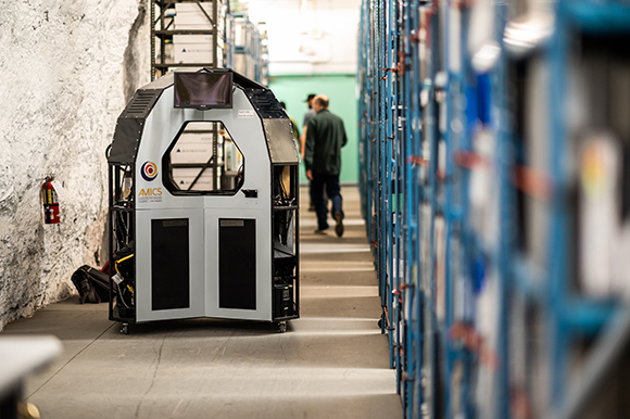 An automated guided vehicle with a screen on top sits in a narrow warehouse aisle next to blue shelving.