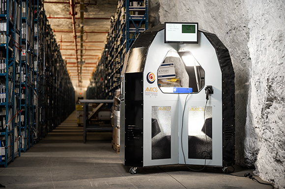 A large black and silver automated machine with a screen stands in a warehouse aisle next to a rocky wall.