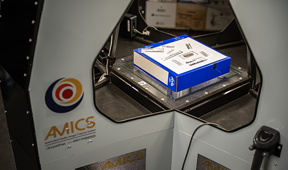 A blue box rests on a glass platform inside a large, grey automated imaging machine labeled AMICS.