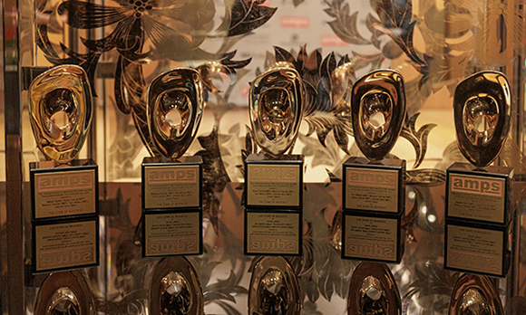 Five gold-colored trophies with black bases arranged in a row on a glass shelf.