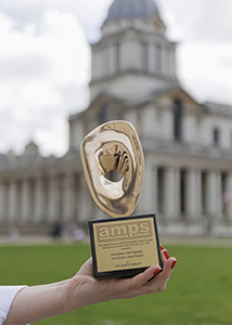 A hand holds a gold, irregularly shaped trophy with a black base in front of a blurred classical building.