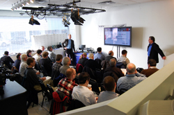 A group of people seated in a room watching a presentation on a large monitor.
